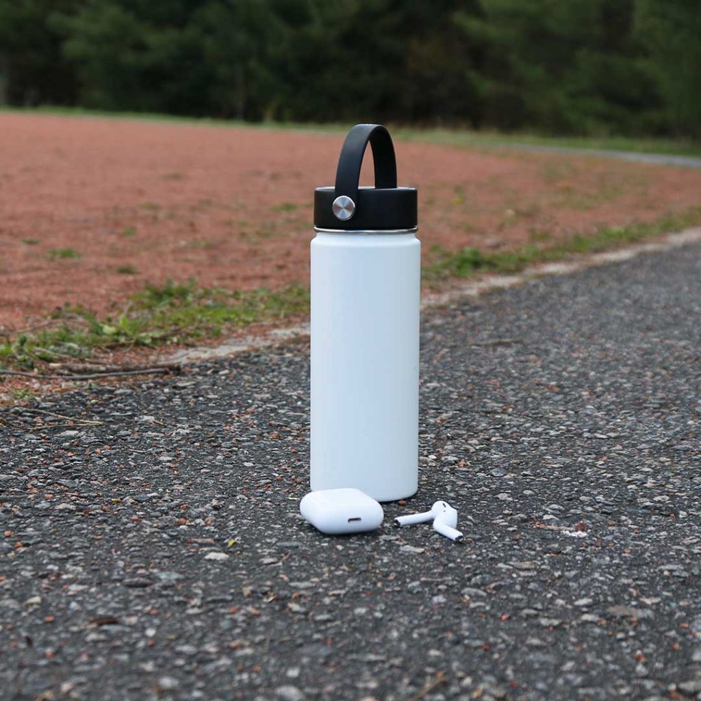 A white metal water bottle with a black lid is placed on the ground of a running track, with a pair of wireless headphones beside it. 