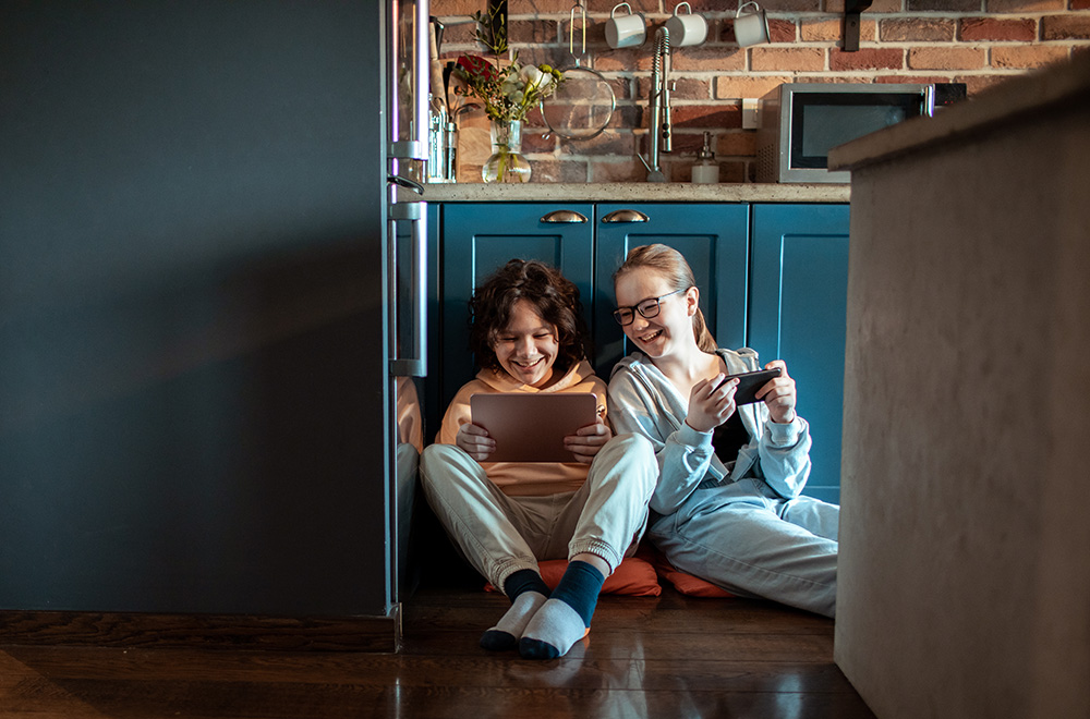 Two young friends sit on kitchen floor smiling at a tablet