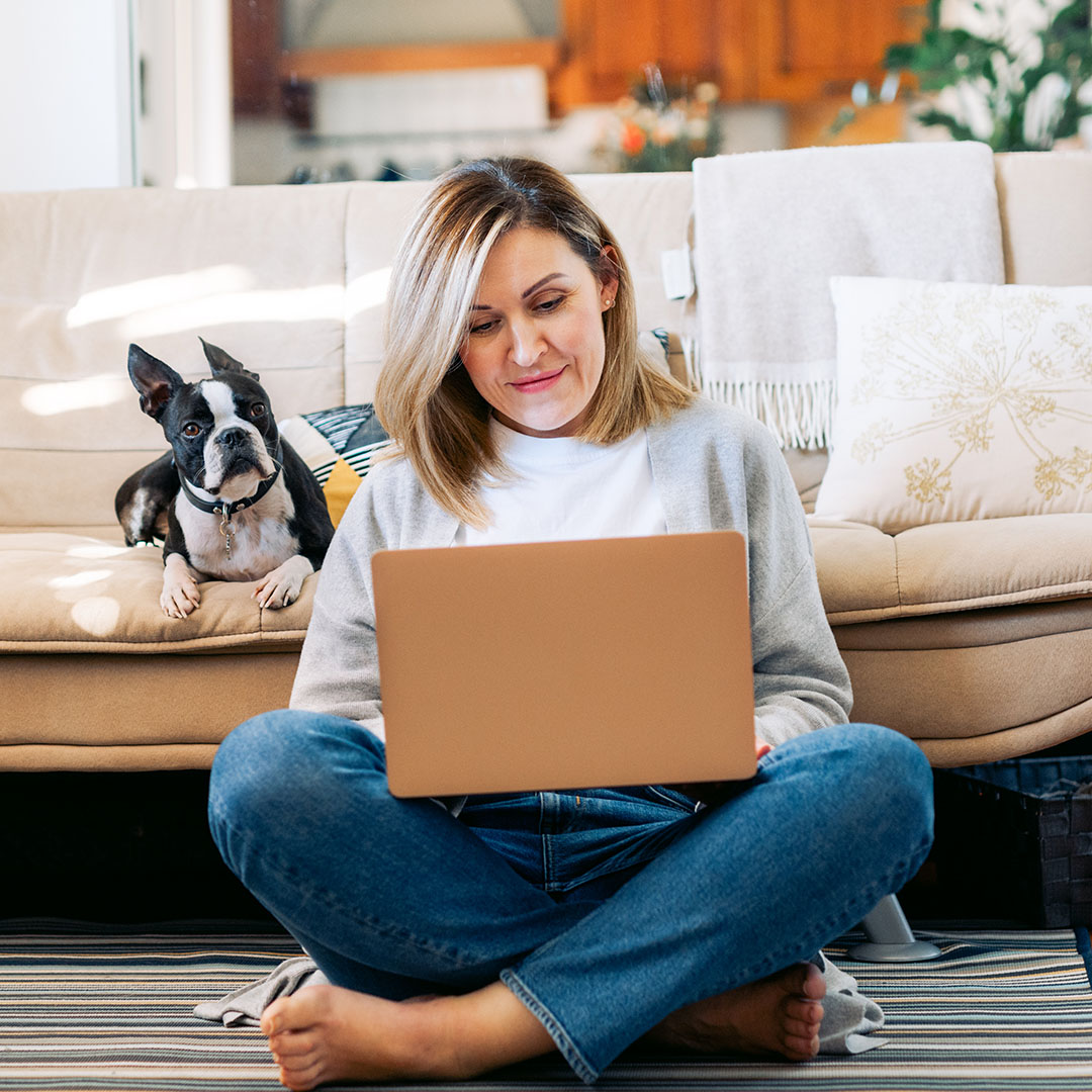 A woman with blonde hair sits cross-legged on the floor with a laptop computer