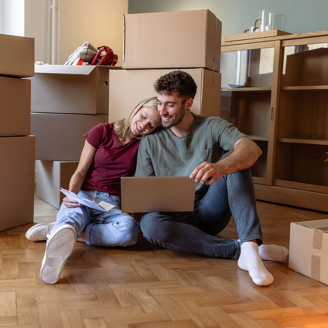 A young couple sits on the floor of their new home with moving boxes in the background, looking at a laptop