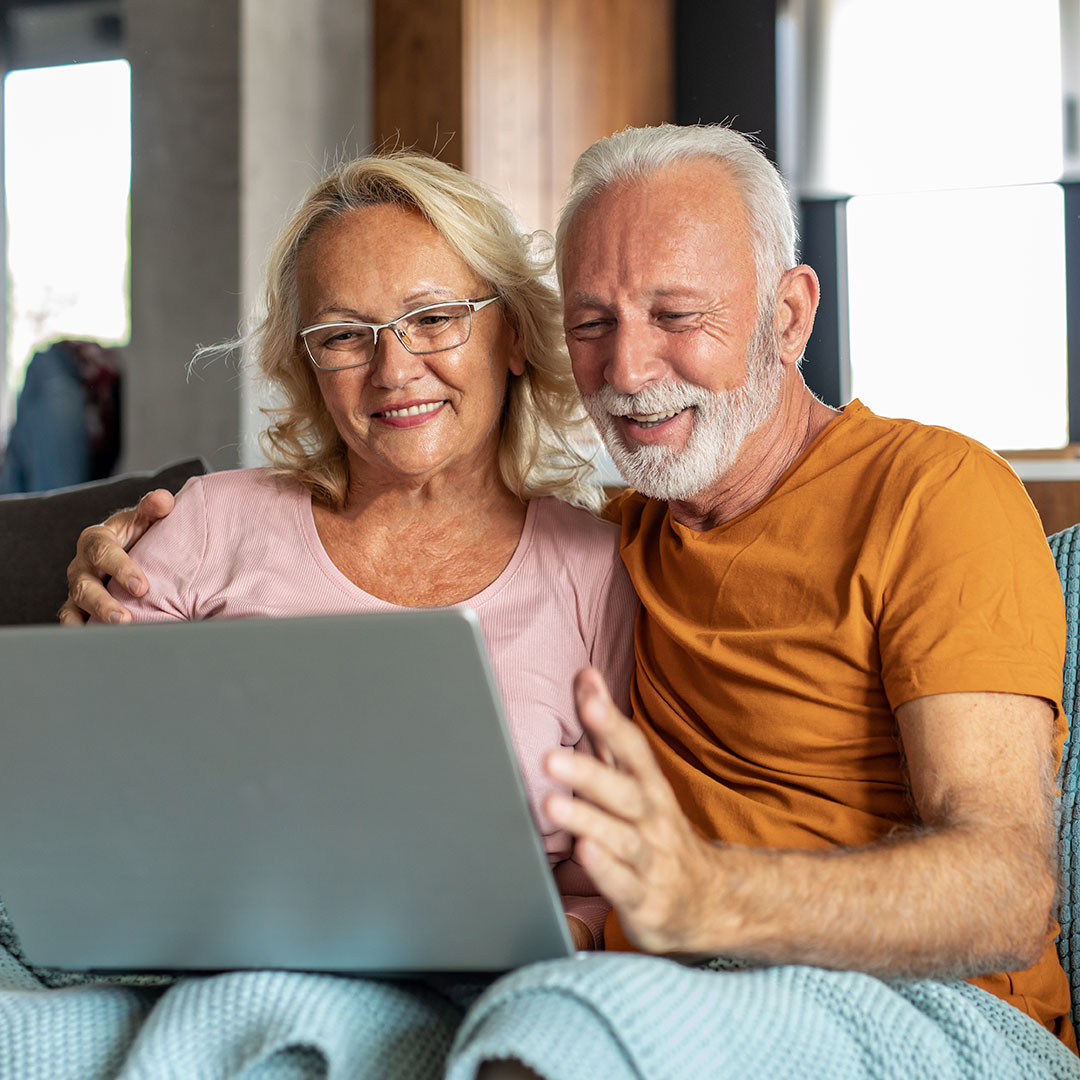 An older man and woman sit on a couch smiling at a computer