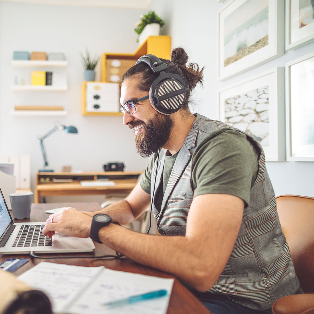 A bearded man wearing headphones sits at a desk and smiles at a computer